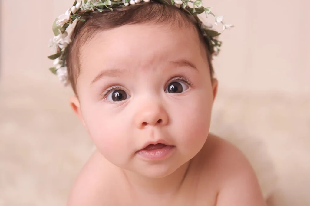 A close-up of a baby with wide brown eyes, light skin, and dark hair, wearing a floral headband and looking curiously at