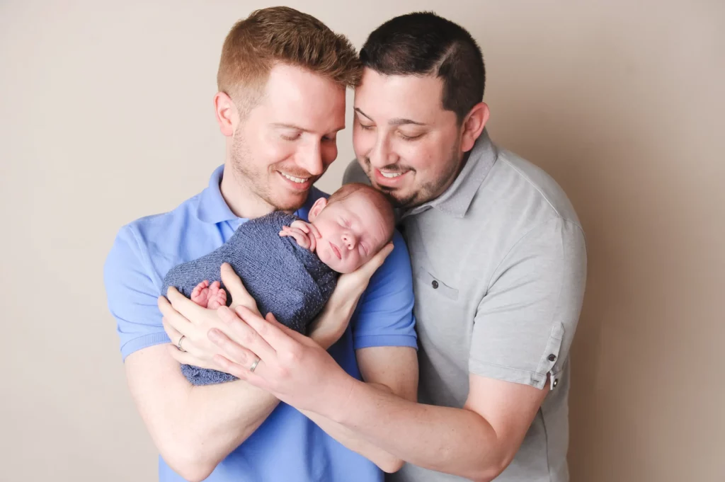 Two smiling men lovingly hold a sleeping newborn wrapped in a blue blanket, standing close together against a neutral
