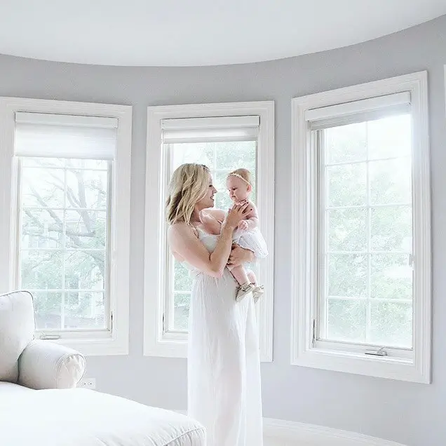 A woman in a white dress holds a baby in a bright, spacious room with large windows