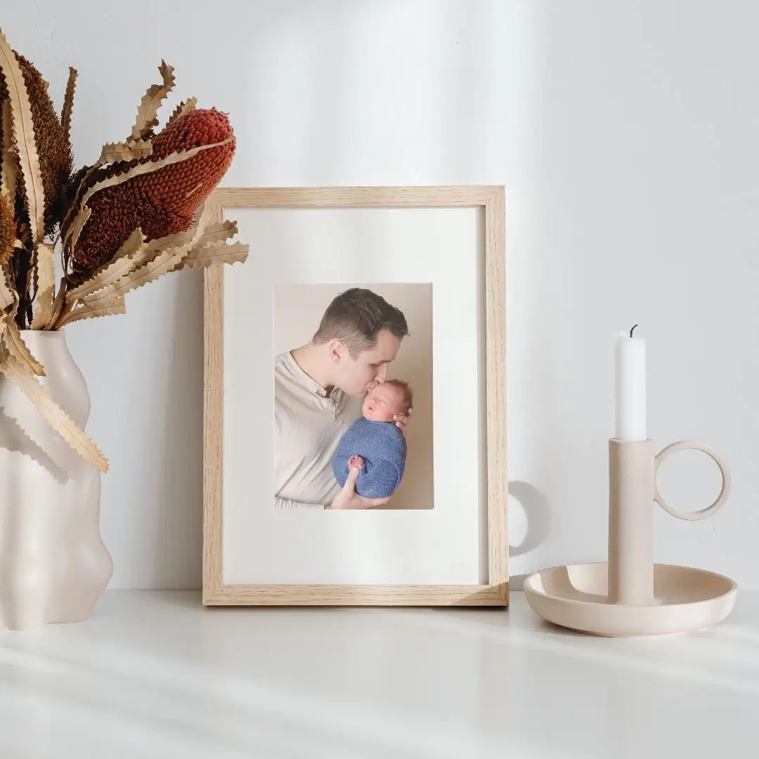 A framed photo of an adult holding and kissing a baby, displayed on a white surface next to a beige candle holder with a
