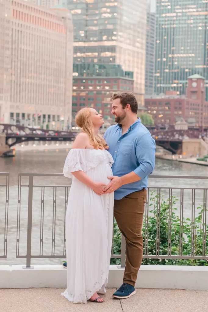 A couple stands in an embrace by a river with city buildings reflecting the light in the background