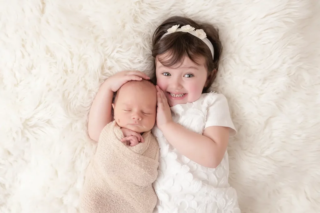 A smiling young girl in a white dress lies on a fluffy white blanket next to a sleeping newborn wrapped in a beige