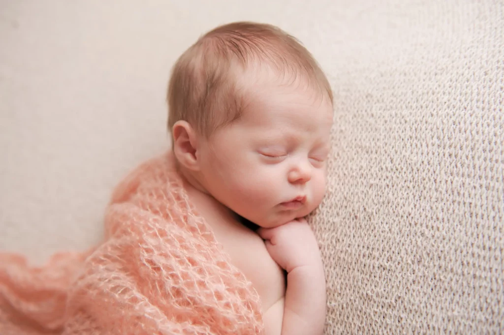 A newborn baby with light hair is peacefully sleeping on a light-textured knit blanket The baby is wrapped in a soft pink