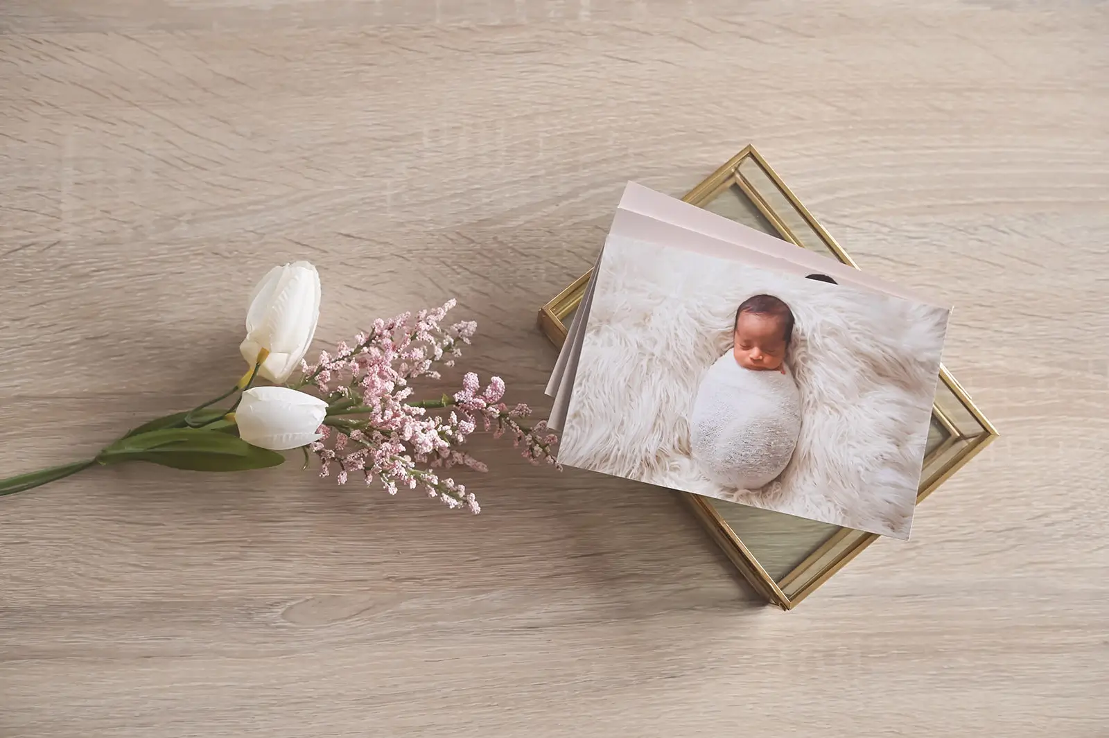 A photo of a sleeping newborn wrapped in a white blanket lies on a glass box, next to two white tulips and pink flowers