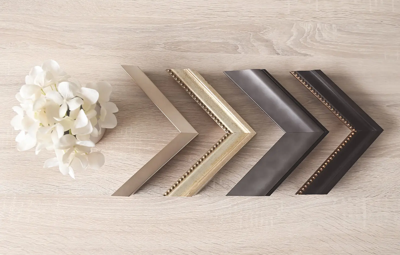 A small bouquet of white flowers sits on a light wooden surface next to five different styles and colors of picture frame