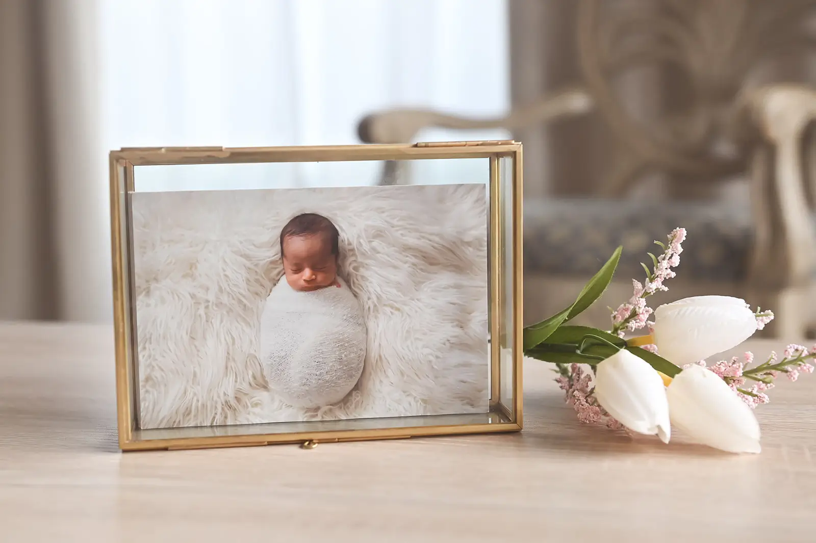 A framed photo of a swaddled newborn baby lying on a soft, white furry blanket sits on a wooden surface next to three
