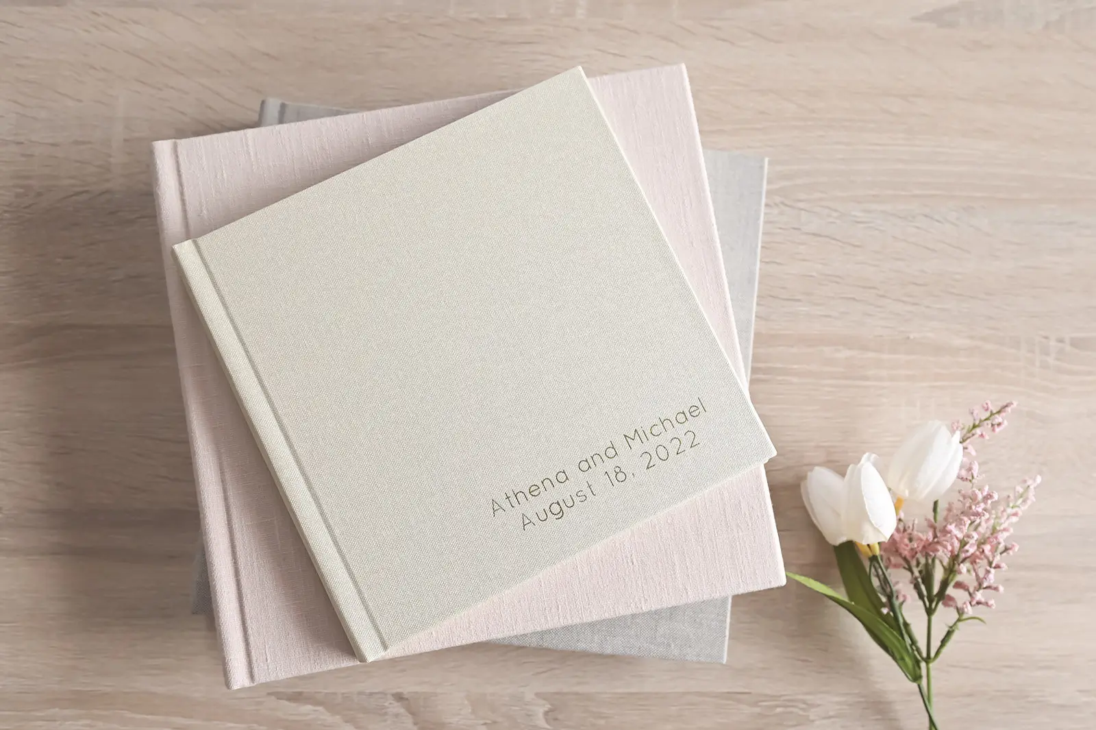 A pale beige photo album with "Athena and Michael August 18, 2022" embossed on the cover rests on top of two closed books